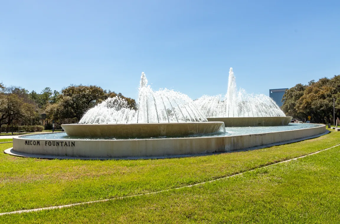 The iconic Mecom Fountain spraying water from its tiered concrete basins, surrounded by a manicured green lawn under a clear blue sky.
