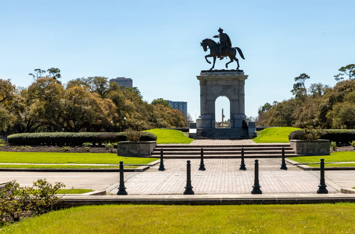 The Sam Houston Monument featuring a bronze equestrian statue atop a large stone arch, situated in a green park with stone walkways and tree-lined paths.