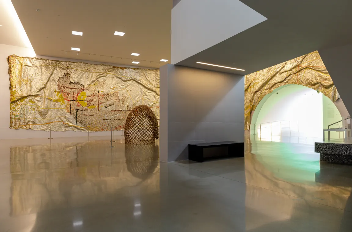 A spacious museum interior featuring a large gold tapestry hanging on the wall, a woven dome structure on the floor, and an arched entryway lit with glowing green light.