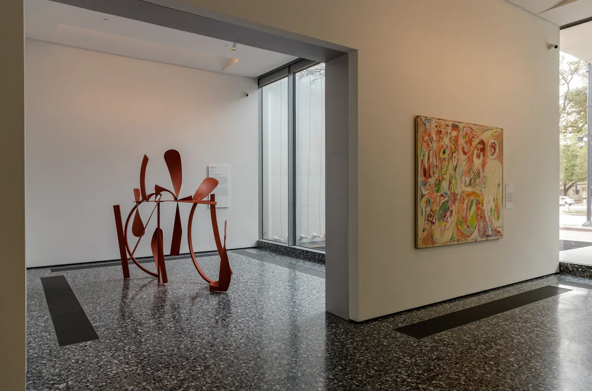A bright gallery space with speckled terrazzo flooring featuring an abstract red metal sculpture and a colorful, textured oil painting hanging on a nearby wall.