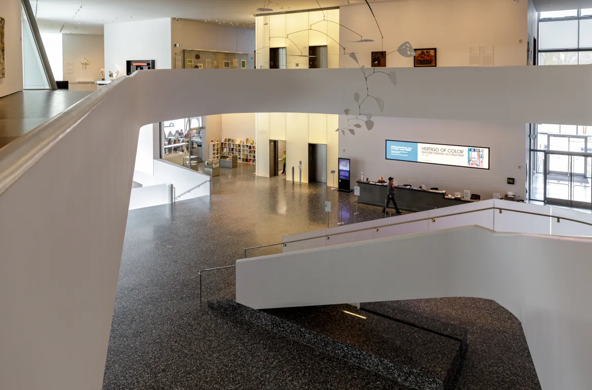 A multi-level museum lobby viewed from above an elevated walkway, showing wide white staircases, a reception desk, and a delicate hanging mobile sculpture.