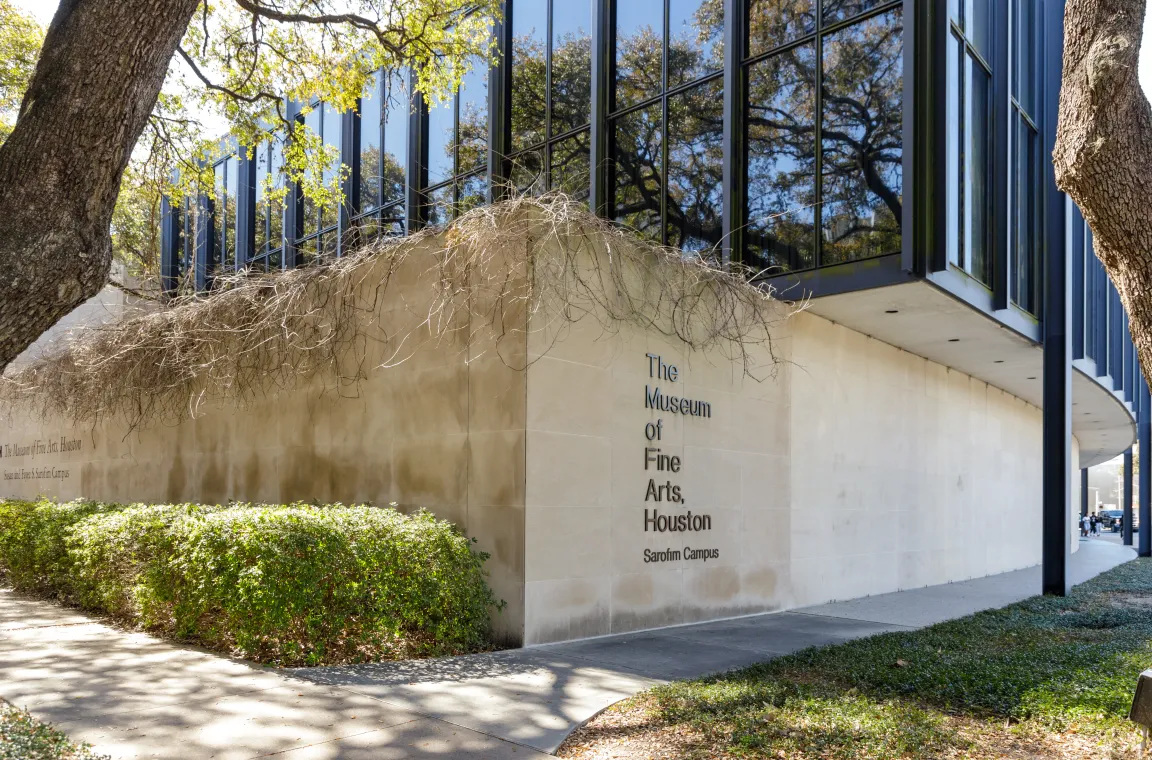 A modern stone building with large glass windows reflecting the surrounding trees, featuring the text "The Museum of Fine Arts, Houston Sarofim Campus" engraved on the wall.