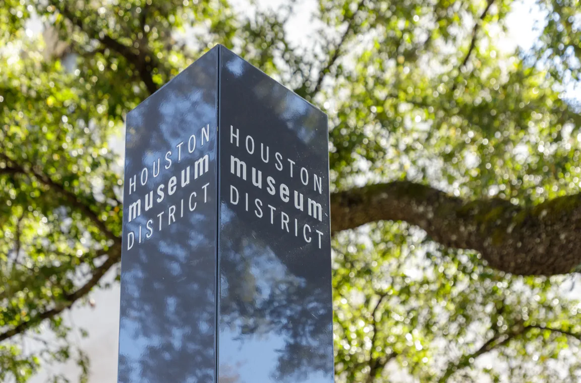 A close-up shot of a glossy black pillar sign that reads "HOUSTON museum DISTRICT" on its sides, positioned under a canopy of green oak trees.