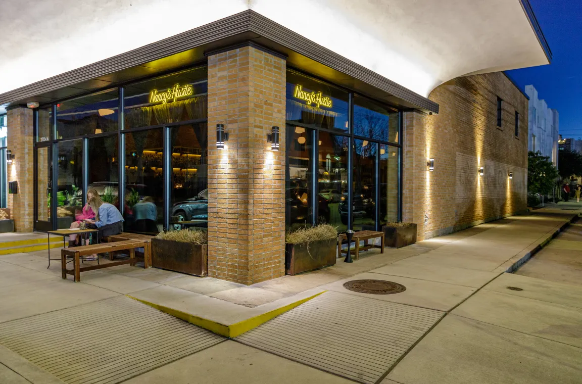An evening exterior shot of a modern brick corner restaurant featuring large glass windows, glowing yellow "Nancy's Hustle" neon signs, and patrons seated at an outdoor wooden bench and table.