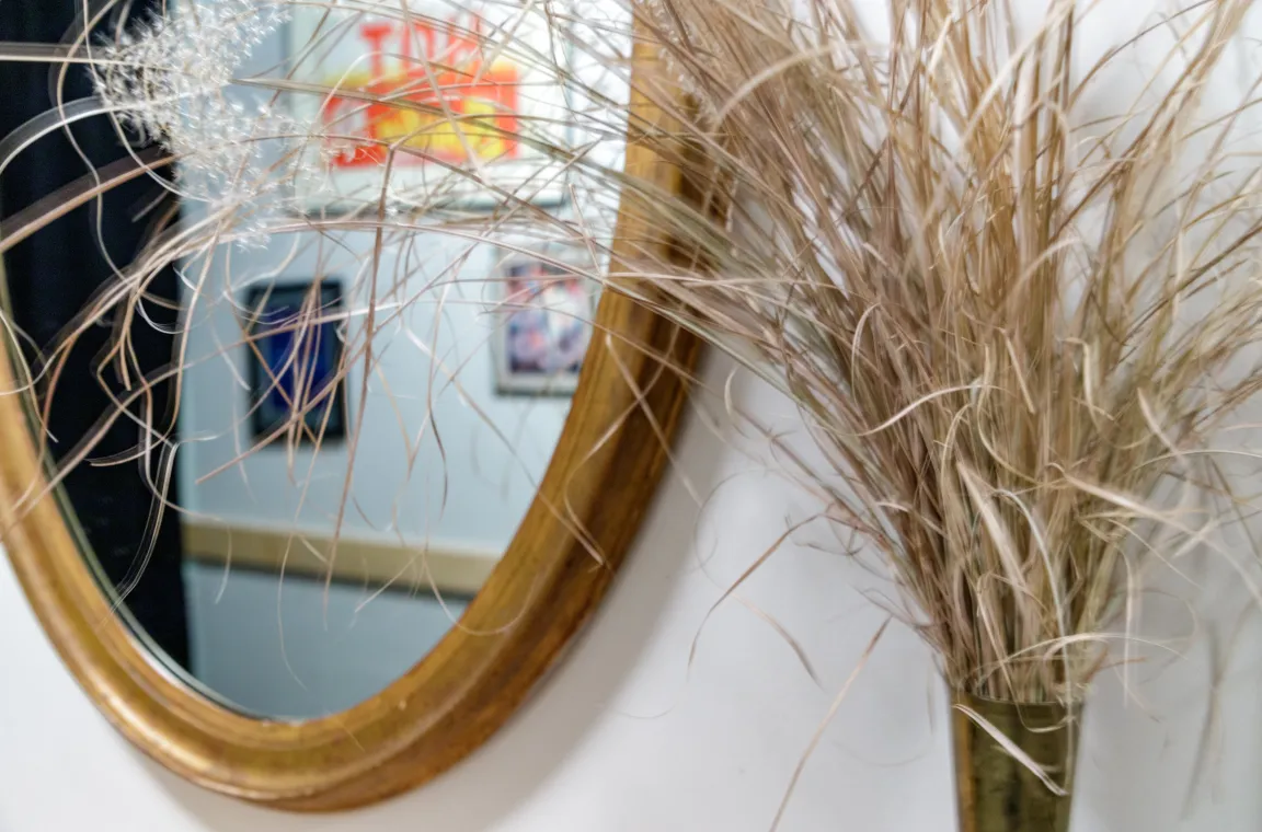 A close-up view of dried decorative grass in a vase, positioned next to a gold-framed oval mirror that reflects blurred framed artwork on an opposite wall.