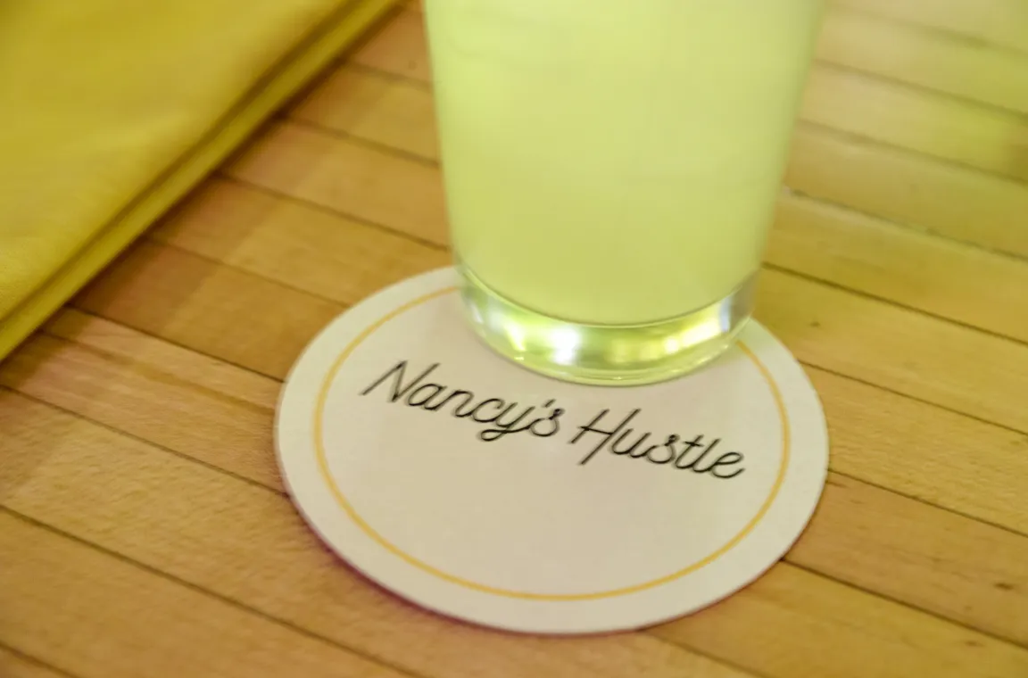 A close-up shot of a round white coaster on a wooden table with the cursive text "Nancy's Hustle" printed inside a thin yellow border, partially supporting a glass of a light green drink.