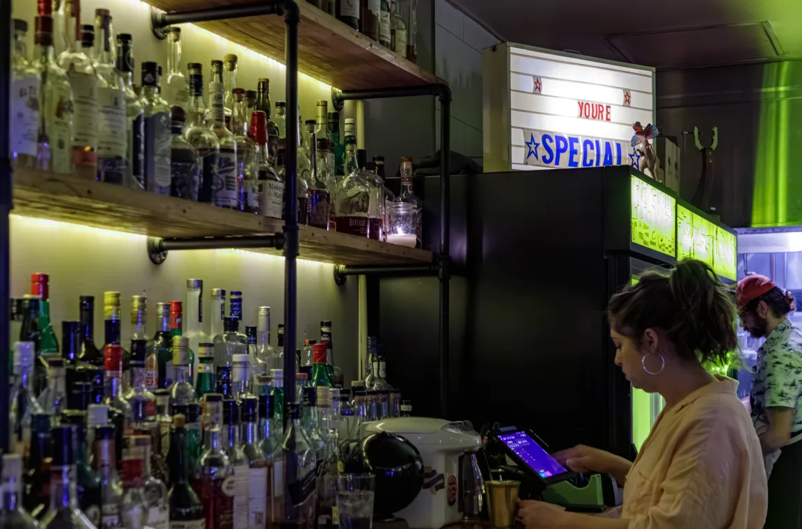 A female bartender working at a well-stocked bar counter under a lighted sign that reads "YOU'RE SPECIAL," with rows of liquor bottles on wooden shelves in the foreground.