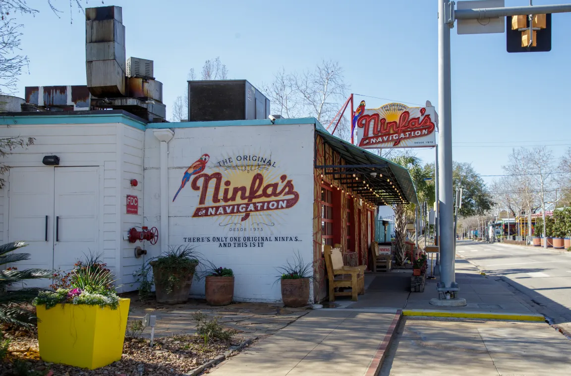 An exterior shot of a white brick restaurant building with a hand-painted mural reading "THE ORIGINAL Ninfa's ON NAVIGATION," featuring an outdoor sidewalk, potted plants, and a rooftop sign.
