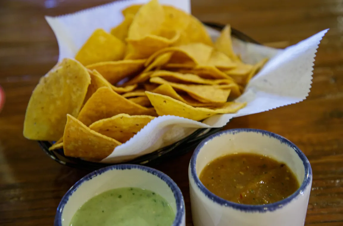 A close-up shot of a wire basket filled with crispy yellow tortilla chips lined with white paper, sitting on a wooden table next to two small bowls of green and brown salsa.