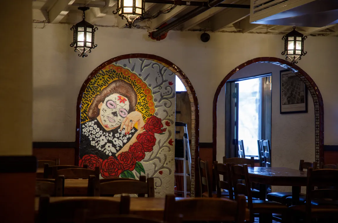 A dimly lit restaurant dining room with dark wooden tables and chairs, featuring hanging lantern lights and a colorful wall mural of a woman with Day of the Dead face paint surrounded by red roses.