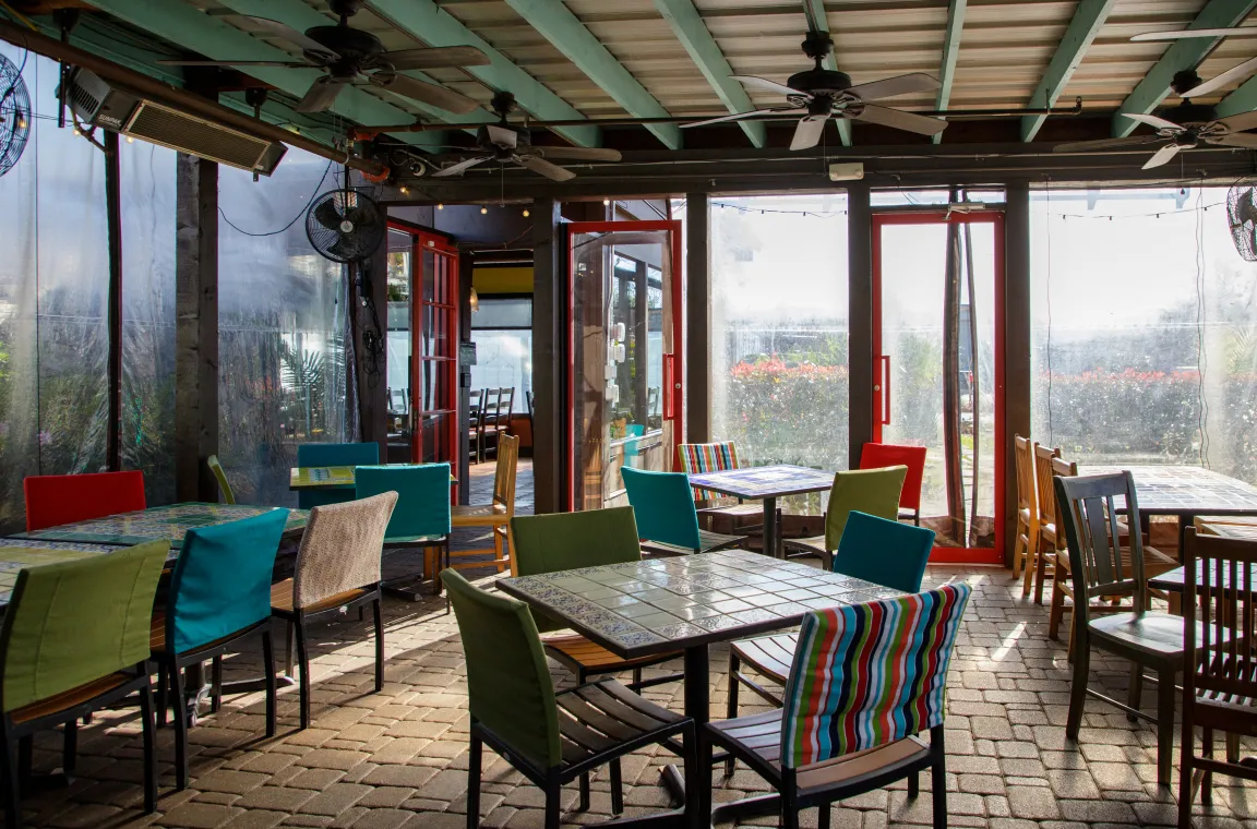 An enclosed patio dining space with a cobblestone floor, multiple square tiled tables, mismatched colorful chairs, and ceiling fans spinning overhead.