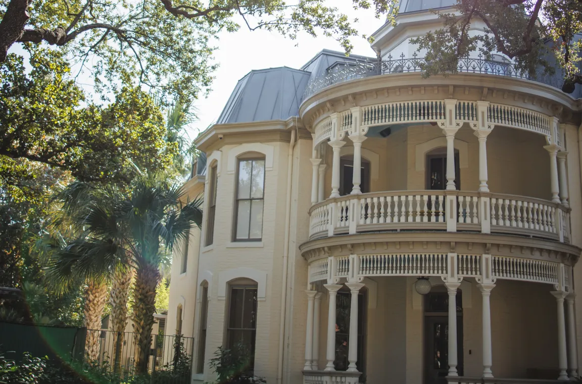 Elegant Victorian mansion with wraparound porches, ornate balconies, and mature oak trees in San Antonio's King William neighborhood.