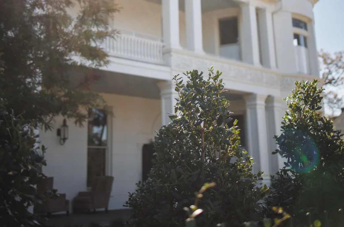 White historic mansion with tall columns and landscaped gardens, photographed in bright afternoon sunlight.