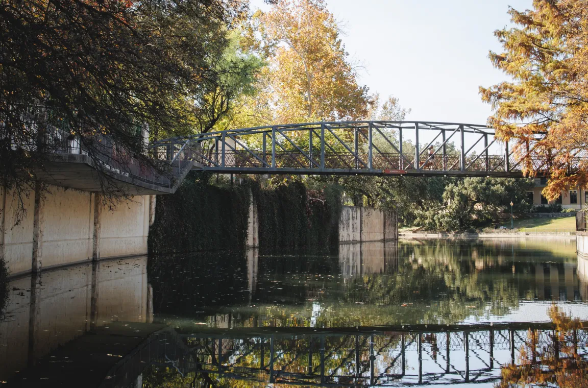 Steel pedestrian bridge crossing the San Antonio River, reflected in calm water beneath autumn-colored trees.