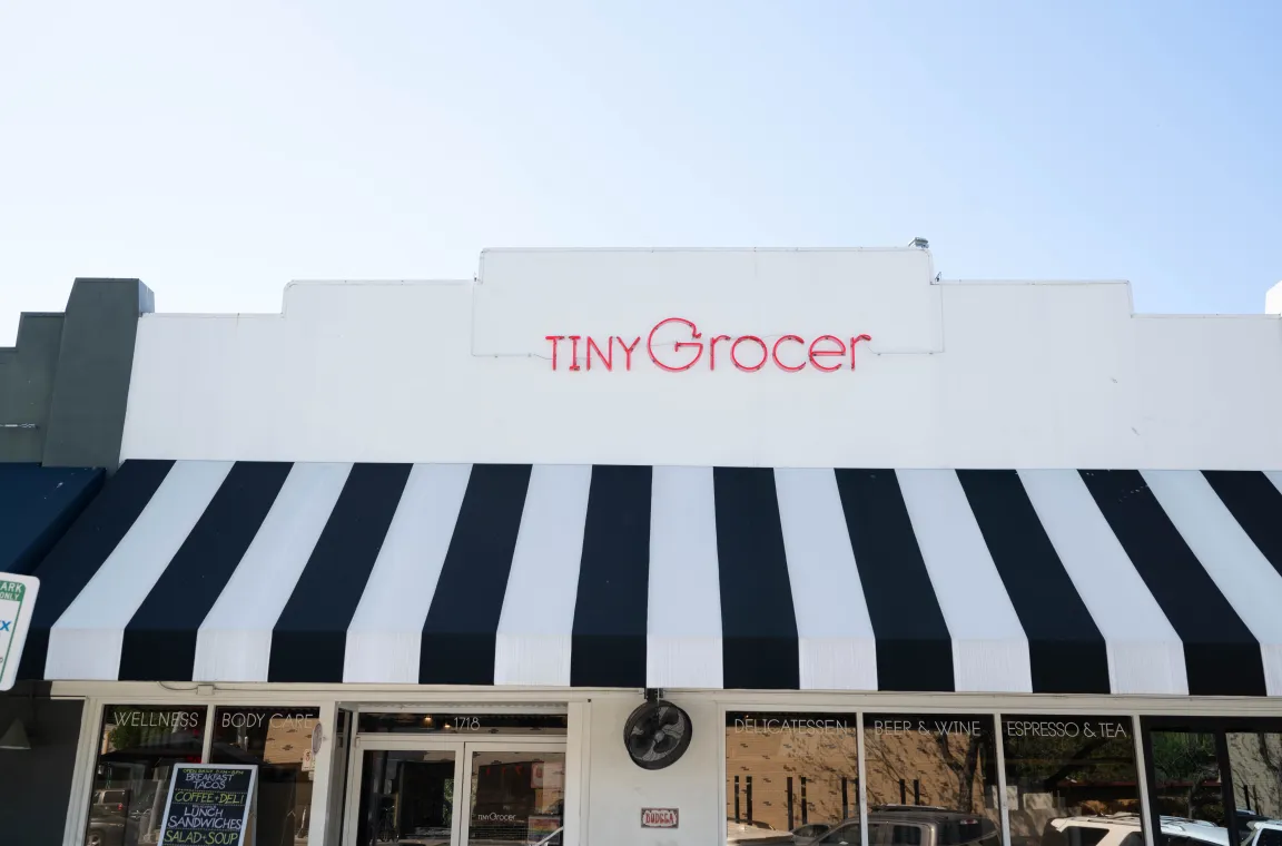 Exterior of Tiny Grocer featuring a black-and-white striped awning and signature red storefront sign.