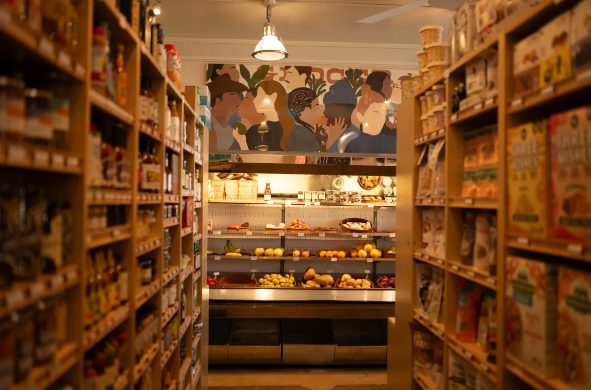 Grocery aisle at Tiny Grocer featuring specialty pantry items, fresh produce, and a colorful mural above the market.