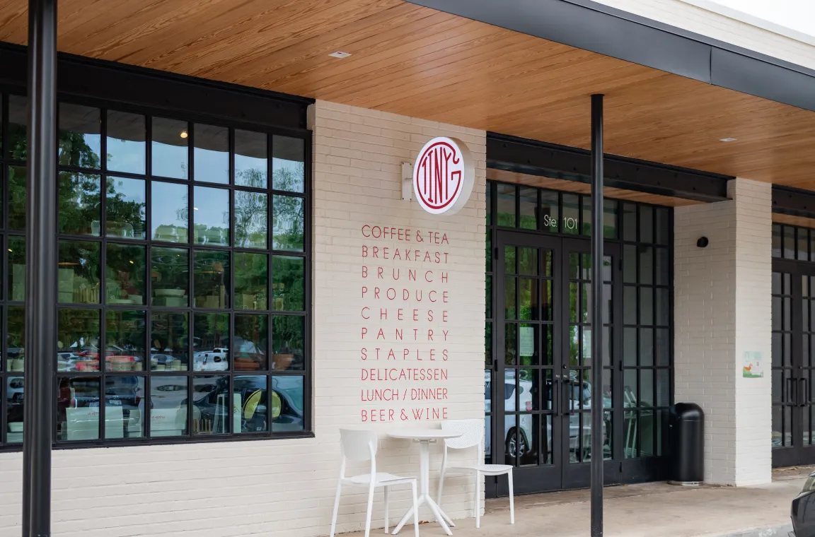 Exterior entrance of Tiny Grocer featuring modern storefront windows, outdoor seating, and a neighborhood market café.