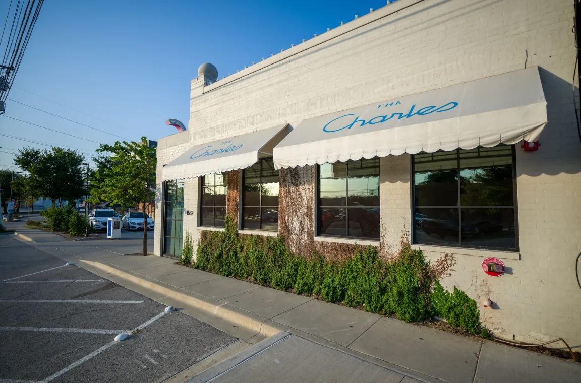 Daytime exterior of The Charles restaurant in Dallas featuring its white brick building, blue awnings, and landscaped facade along Knox Street.