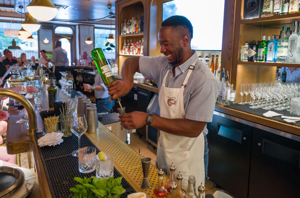 Bartender preparing a cocktail behind the bar at Green Point Seafood & Oyster Bar in Dallas, with bottles, glassware, and guests visible throughout the lively dining room.