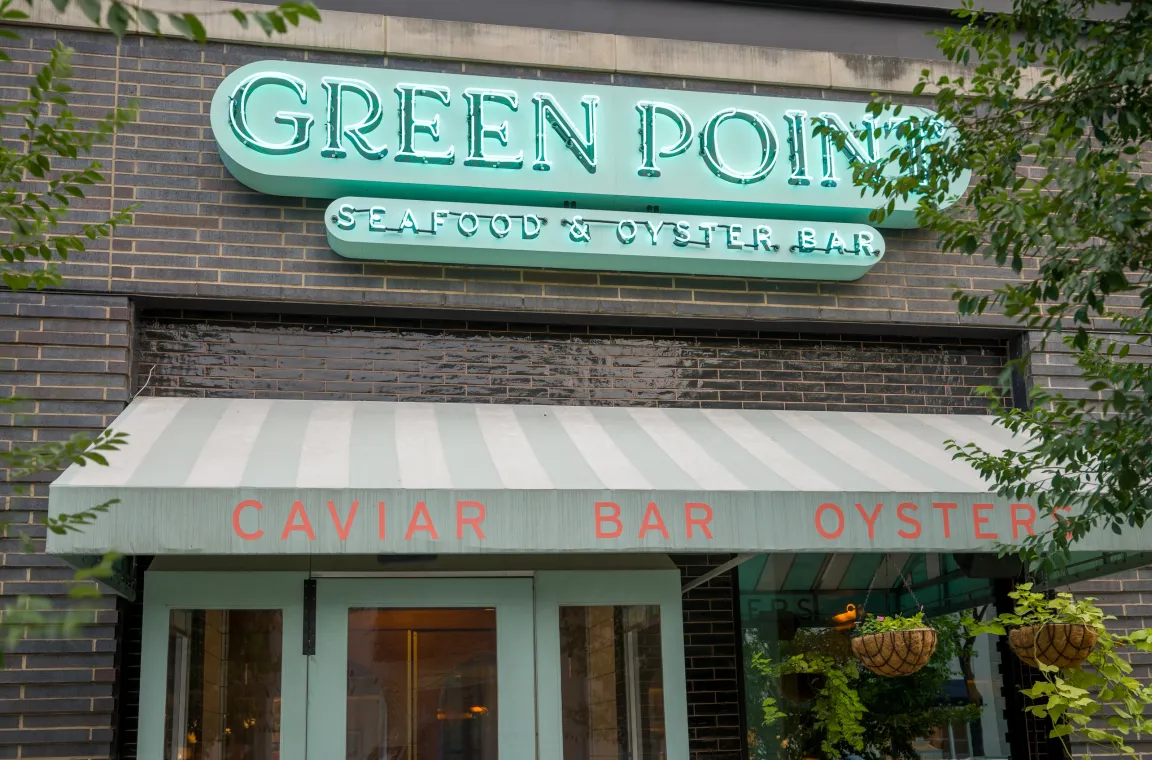 Exterior entrance of Green Point Seafood & Oyster Bar in Dallas featuring its illuminated sign above a striped awning with caviar bar and oyster bar branding.