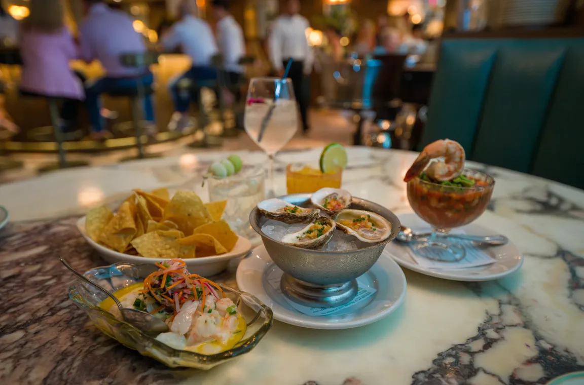 Assorted seafood appetizers, including oysters on the half shell, shrimp cocktail, ceviche, and tortilla chips, served with cocktails on a marble table inside Green Point Seafood & Oyster Bar.