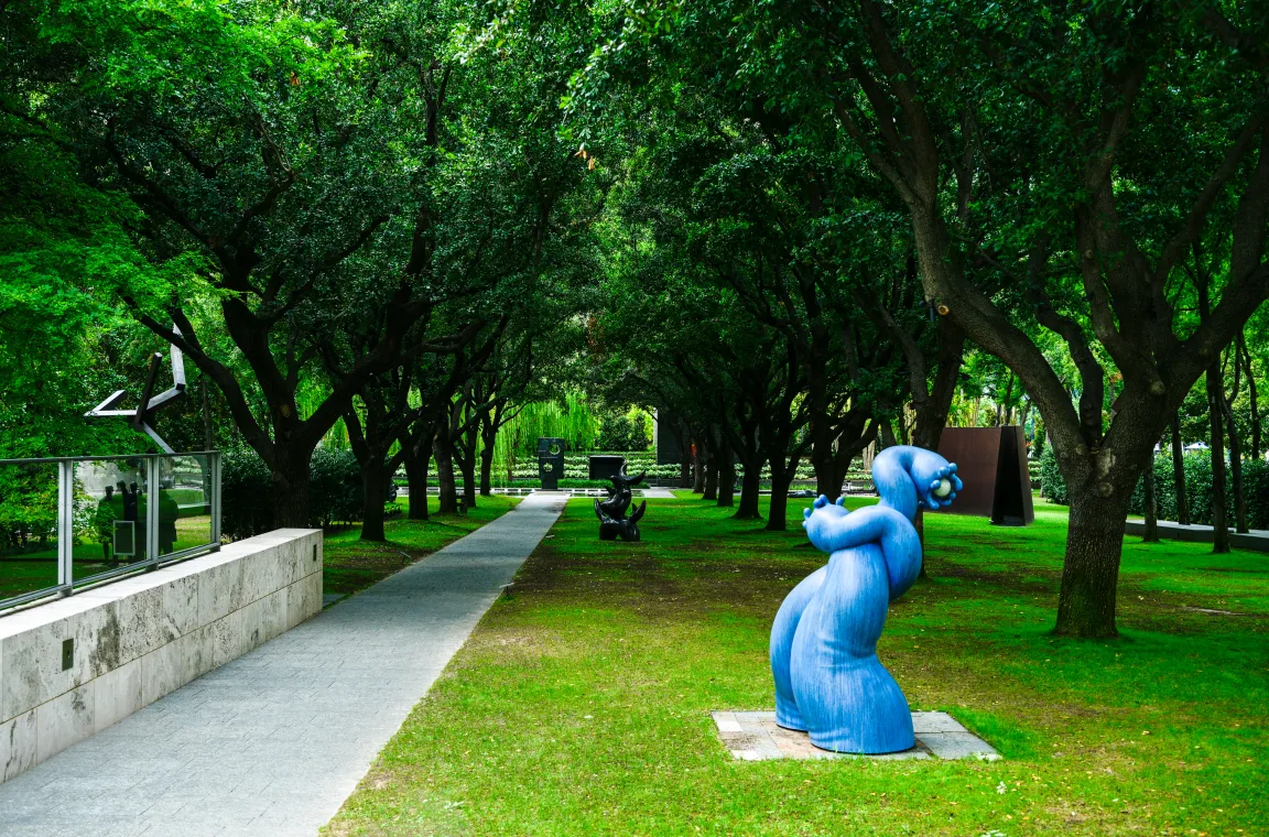 The Nasher Sculpture Garden's shaded oak-lined pathways featuring contemporary sculptures integrated into the landscape, including a striking blue abstract figure in the foreground.