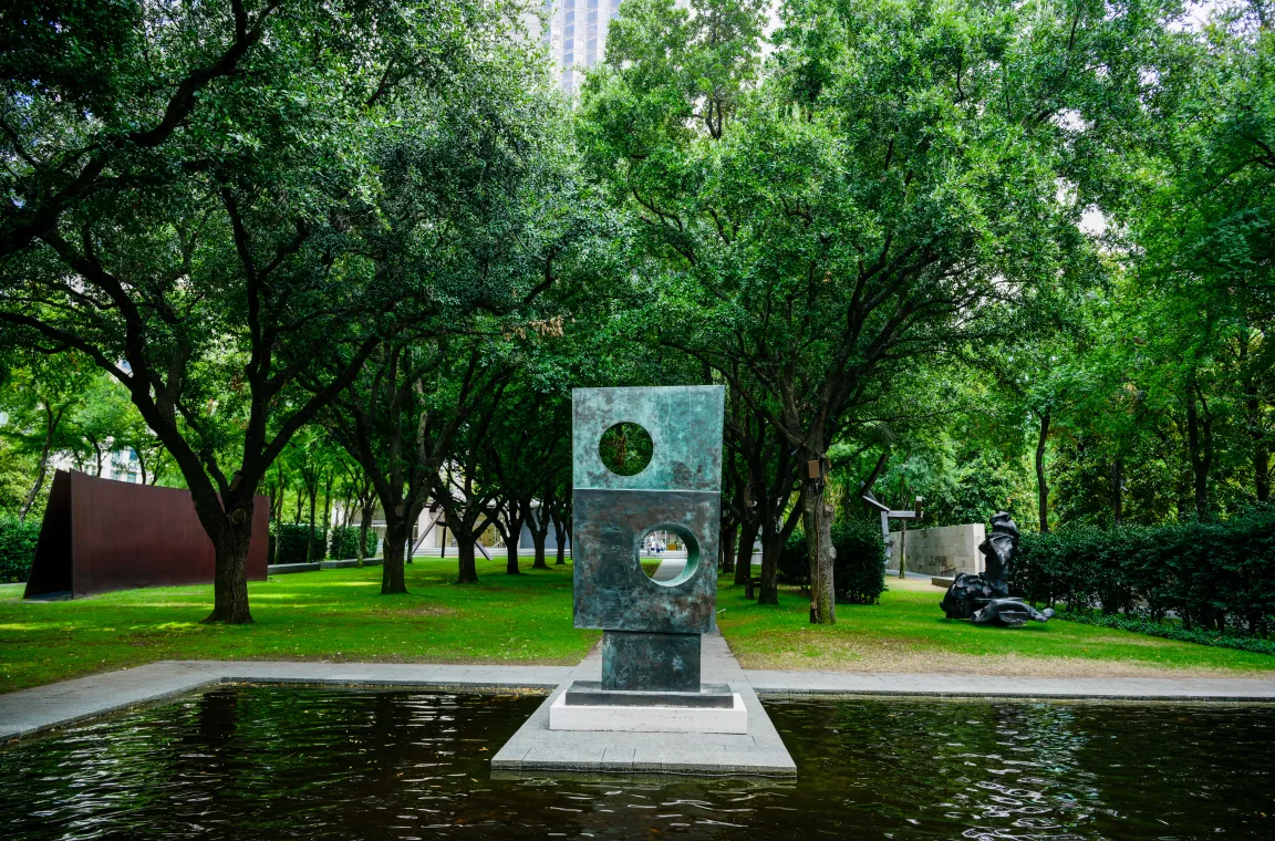 A monumental bronze geometric sculpture reflected in a shallow pool, framed by mature trees and the Sculpture Garden's carefully designed outdoor galleries.