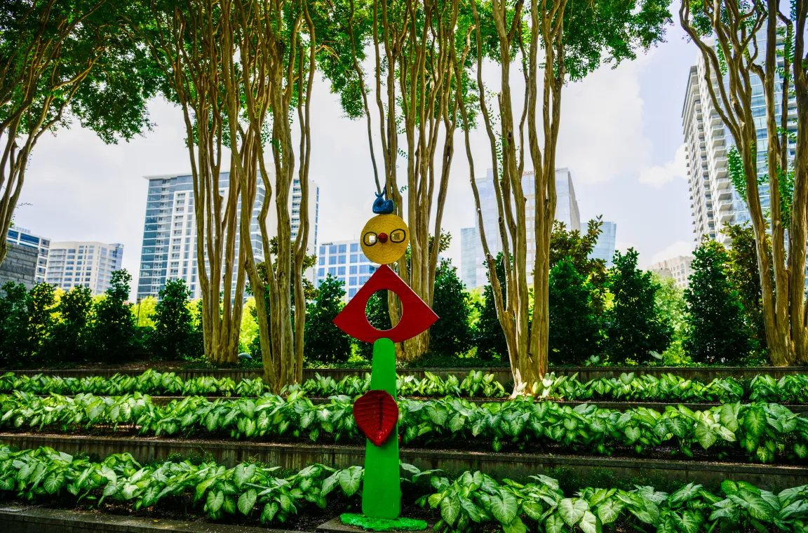 A colorful contemporary sculpture set among terraced gardens with the Dallas skyline rising beyond the tree canopy, highlighting the museum's unique urban setting.