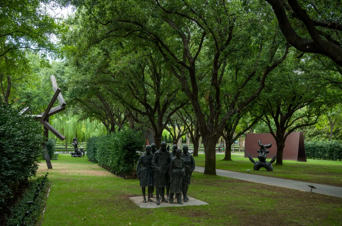 An outdoor gallery featuring a group of bronze figures alongside monumental abstract sculptures beneath a canopy of mature live oaks, showcasing the diversity of the Nasher's permanent collection.