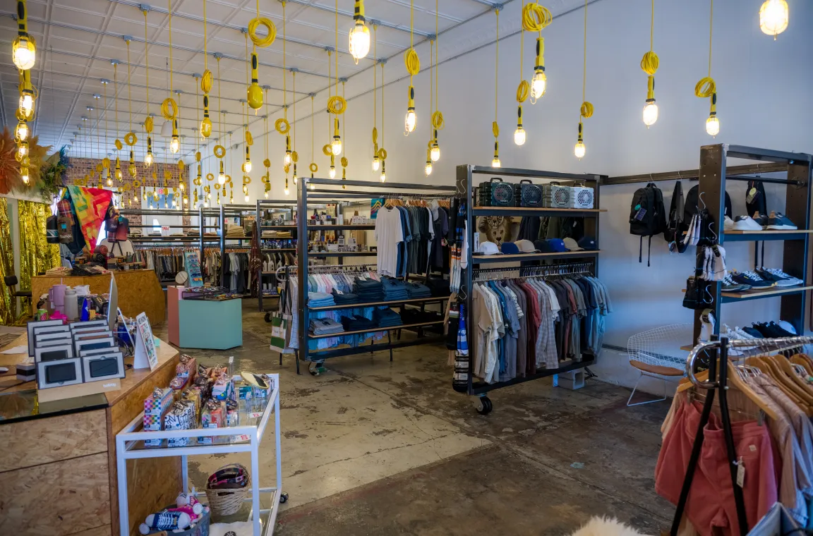 Interior of DLM Supply boutique in Dallas, featuring industrial-style shelving with apparel, accessories, and footwear beneath rows of hanging yellow work lights.