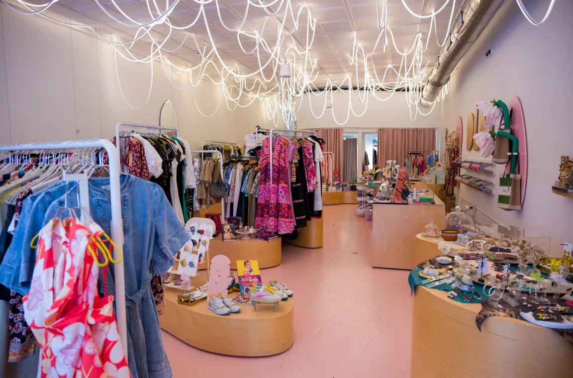 Bright interior of DLM Supply showcasing colorful women's clothing, jewelry, and gifts beneath a sculptural ceiling installation of looping LED light fixtures.