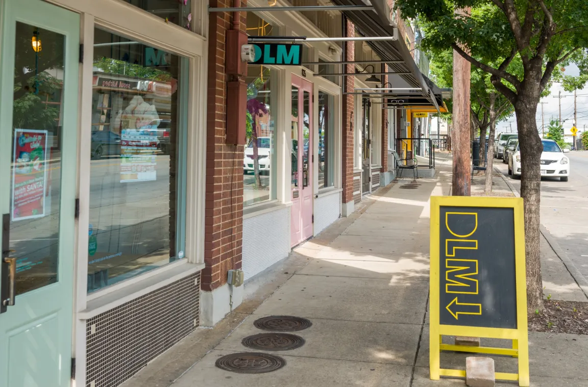 Sidewalk view of DLM Supply's storefront in the Bishop Arts District, with the boutique's pink entrance, storefront signage, and neighboring local businesses along the tree-lined street.