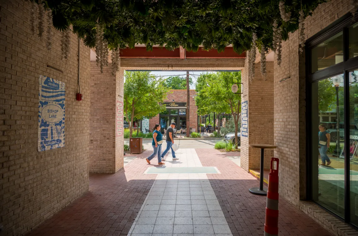 A covered pedestrian passage framed by brick walls and hanging greenery, opening onto the walkable streets of the Bishop Arts District with local shops and visitors beyond.