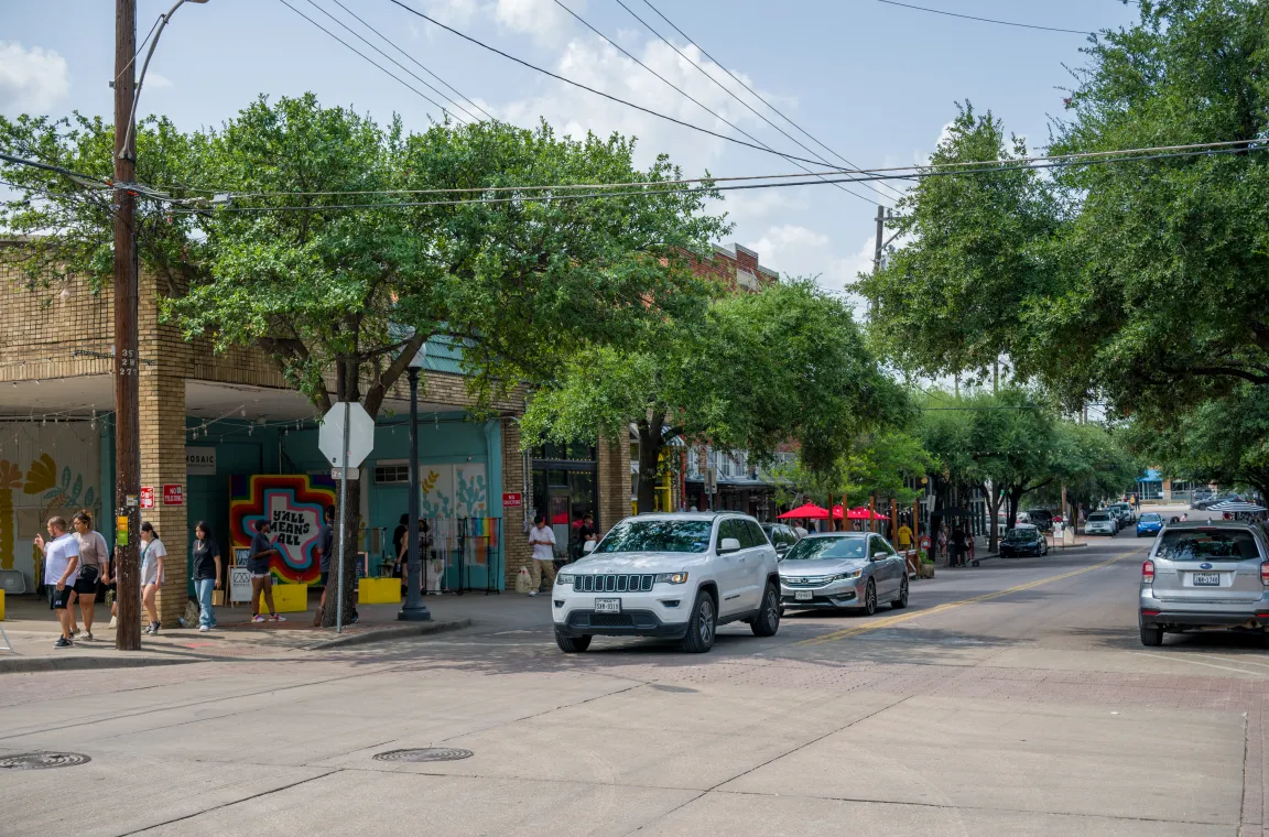 A streetscape along Bishop Arts District lined with locally owned shops, colorful storefronts, mature trees, and pedestrian-friendly sidewalks that define the neighborhood's eclectic character.