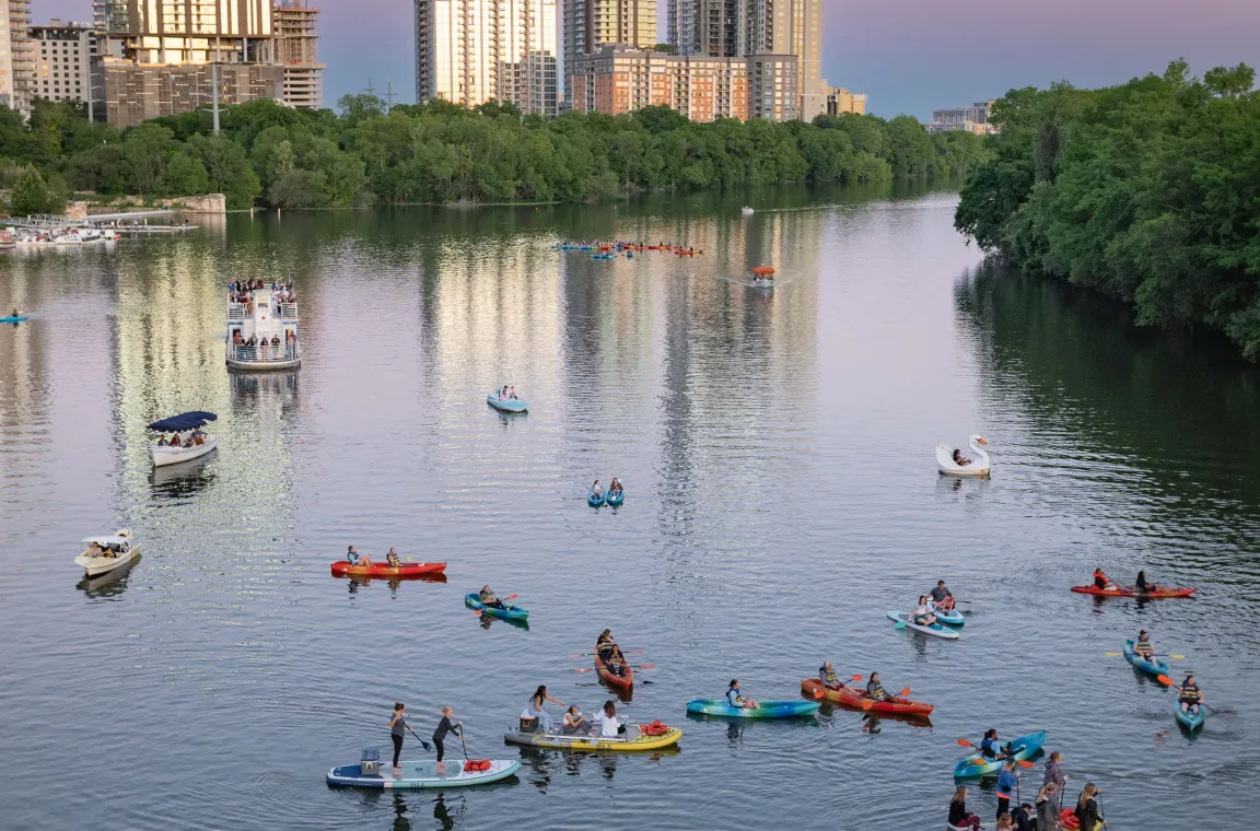 Twilight view of Lady Bird Lake filled with kayakers, paddleboarders, and boats, with the Austin skyline reflecting on the calm water.