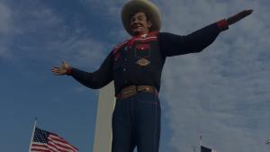 Big Tex, the iconic 55-foot-tall cowboy statue at the State Fair of Texas, standing with outstretched arms beneath a blue Texas sky with American and Texas flags waving below.